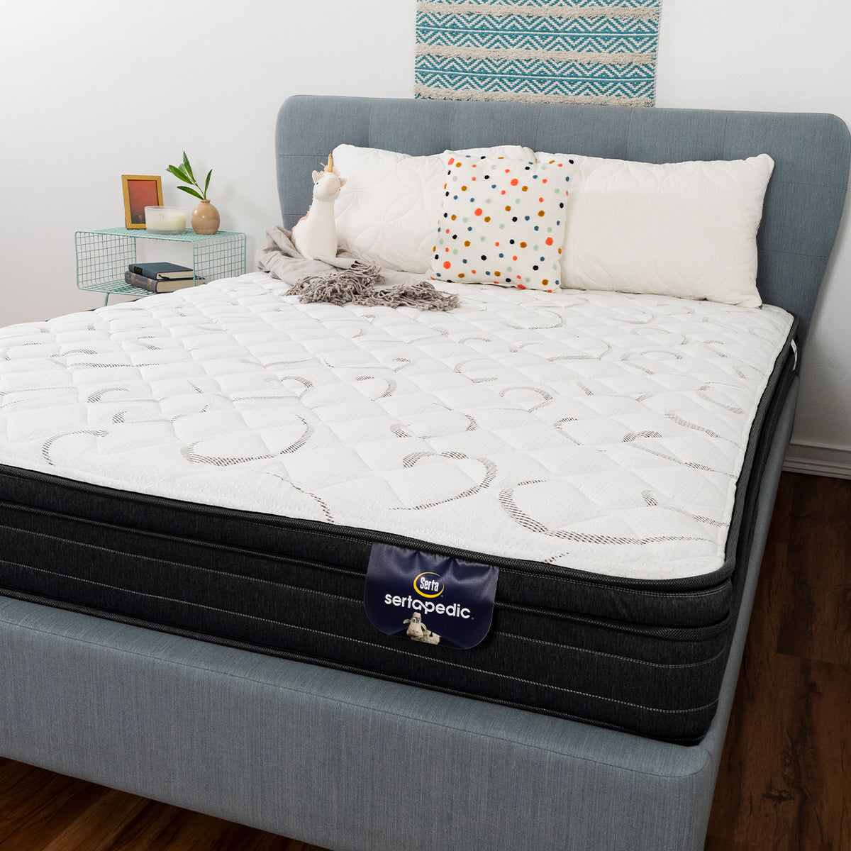 Sertapedic® Elkridge Euro Top Mattress by Serta With Blank, Unicorn Plushie, And Pillows On Bed Frame In Bedroom With A Bedside Table. Front View Showing Product Tag Label And Fabric Detail.