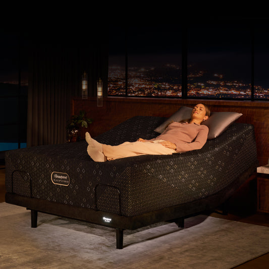 Beautyrest Black Luxury Adjustable Bed Base showing a woman reclines on a raised bed with a patterned mattress, set in a dark bedroom with a city skyline visible at night through the window.